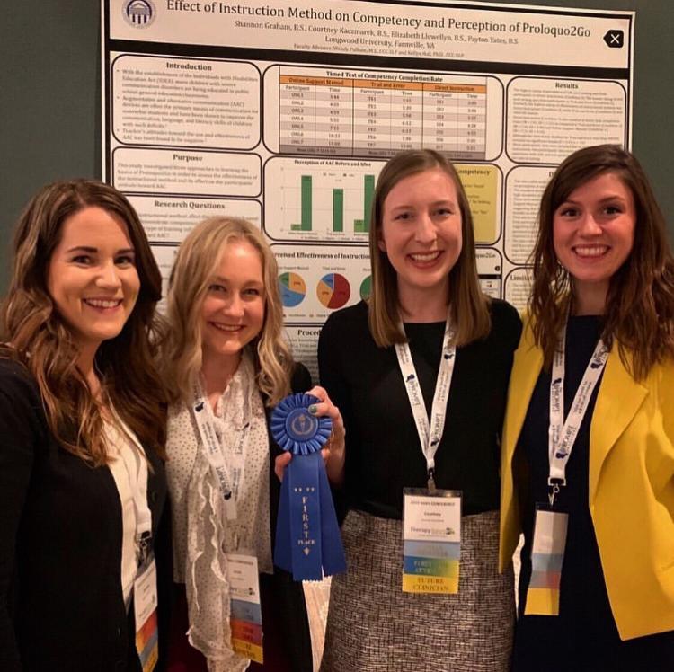 - Four women stand smiling in front of a research poster. One holds a blue first-place ribbon toward the camera. They wear conference badges and professional attire. Charts, graphs, and text from their project fill the poster behind them.