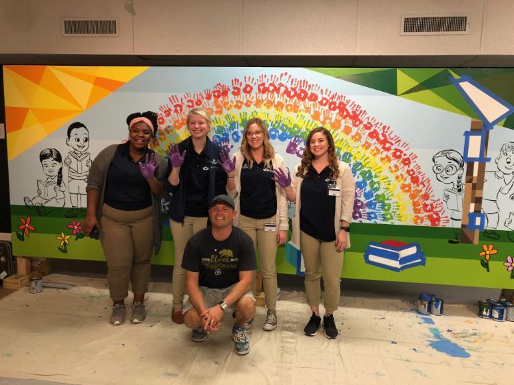 - Five people pose in front of a brightly painted mural with a rainbow of handprints. Four stand smiling with purple paint on their hands while one kneels in front. They wear matching navy shirts and khaki pants in a paint-splattered workspace. Cartoon children, books, flowers, and bold colors fill the cheerful mural behind them.