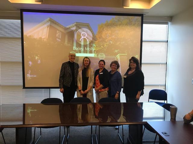 Thesis Completion  - Five people stand together in front of a large projection screen. The screen shows a Longwood University login image with “Dailey, Molly” displayed. They are posed in a conference room with a long reflective table. Bright window light fills the room behind them.
