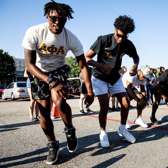 - A group of students dances energetically outdoors in the sunlight. Two students in the front wear fraternity shirts and sunglasses as they step in sync. More students move behind them, joining the lively routine. Cars and campus buildings sit in the background under a clear blue sky.