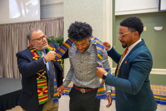 Donning of the Kente is a rite of passage recognizing seniors who have had an impact on diversity, equity, and inclusion at Longwood - Two men place a colorful Kente stole around a student’s shoulders during a ceremony. The student looks down with a smile as the stole is adjusted. All three are dressed in formal or semi-formal attire in a decorated indoor venue. A table covered with additional Ken
