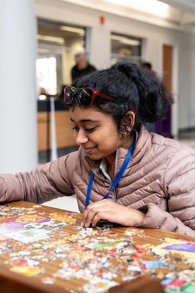 A student wearing a light brown jacket and colorful sunglasses on her head concentrates while assembling a jigsaw puzzle spread across a wooden table.