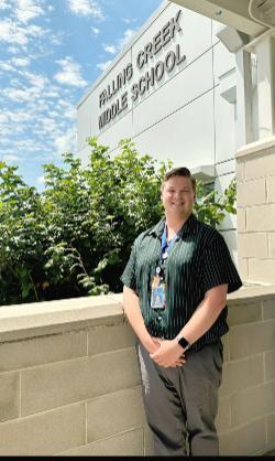 - A person stands smiling outside a school building beneath a sign that reads “Falling Creek Middle School,” with bright greenery and a blue sky with scattered clouds in the background.