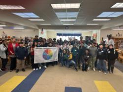- A large group of middle-school students and staff gather in a school library, smiling for a group photo. Several students in the front hold a banner featuring a colorful “Sources of Strength” wheel. The room is bright with bookshelves, posters, and string lights in the background.