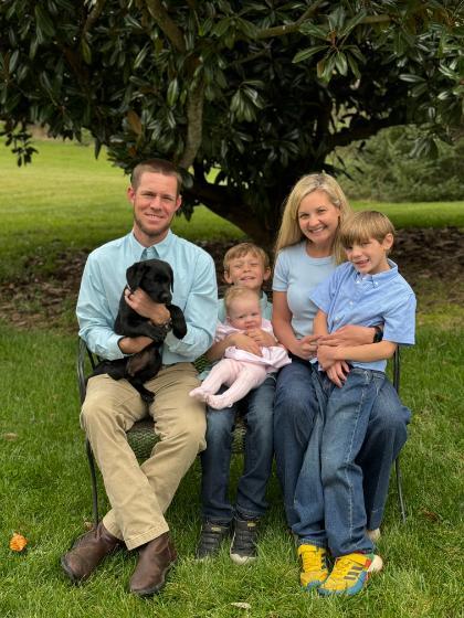 - A family of five sits together on an outdoor bench beneath a large magnolia tree. The father holds a small black puppy on his lap, while two young boys smile beside their parents. The mother holds a baby dressed in pink, and the family is casually dressed in light blue and denim. Green grass and leafy branches create a warm, natural backdrop.