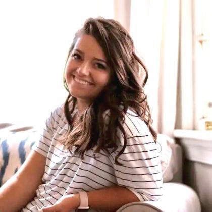 A woman with long, wavy brown hair sits on a couch, smiling warmly at the camera. She is wearing a white shirt with thin black stripes and a smartwatch on her wrist. The background features soft lighting, white curtains, and a cozy living room setting.