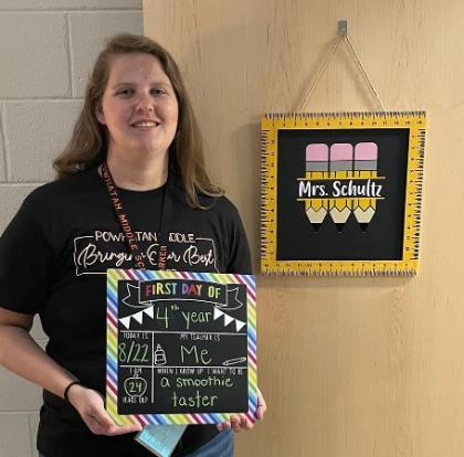 Erin Shultz holds a first day of school sign - A woman stands smiling in a school hallway holding a colorful “First Day Of” sign. She wears a black Powhatan Middle School T-shirt and a lanyard. The sign lists fun first-day details, including “4th year” and “When I grow up I want to be a smoothie taster.” Behind her, a classroom door displays a framed pencil-themed sign that reads “Mrs. Schultz.”