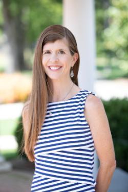 Vertical of Dorothy Suskind - A woman with long straight hair smiles while standing outdoors. She wears a navy-and-white striped sleeveless top with a delicate necklace and patterned drop earrings. Soft natural light brightens her face, and blurred greenery fills the background. She appears relaxed and cheerful in this professional portrait.