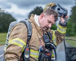Firefighter in turnout gear and breathing apparatus lifts his helmet after responding to an emergency, looking tired but composed.