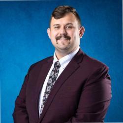 Man in a maroon suit jacket, patterned tie, and checkered shirt, smiling in front of a bright blue background.