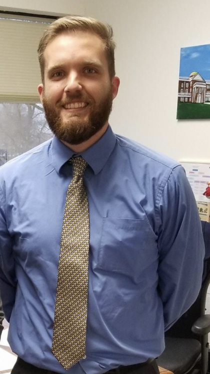 Joseph (Joey) Siverd BS'15, MS'18, Longwood's magic is the faculty. - A man stands indoors wearing a blue dress shirt and patterned tie. He has short light brown hair, a full beard, and a friendly smile. A window and office materials are visible behind him. A colorful painting of a building hangs on the wall to his right.