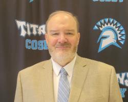 A middle-aged man with light skin, a short beard, and short-cropped hair stands in front of a black backdrop featuring a blue and white Spartan-style helmet logo and the words 