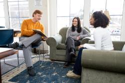 Dr.Justin Jordan talks to two students and shows them a clip board. - A man sits in a blue chair with a clipboard, talking with two students seated on green armchairs in a bright lounge area. Large windows behind them let in natural light and show a view of campus. All three are smiling and engaged in conversation. A patterned rug and wooden tables add warmth to the comfortable study space.