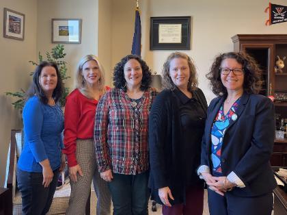 - Five women stand together in an office, smiling at the camera. They are dressed in professional attire in blue, red, plaid, black, and a navy blazer. Framed certificates, photos, and a UVA basketball pennant decorate the wall behind them. The setting includes office furniture, shelving, and soft natural light.