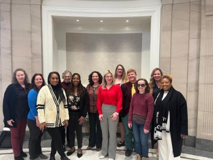 - A group of twelve people stand together in a marble-lined hallway, smiling at the camera. They wear a mix of professional and casual clothing, including sweaters, blazers, and a bright red blouse. The setting features tall stone walls, decorative molding, and soft overhead lighting. The group is arranged in two rows, creating a warm, friendly, and organized portrait.