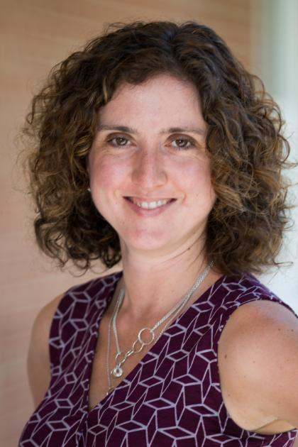 Kat Alves - A woman with short, curly brown hair smiles at the camera in soft, natural light. She wears a sleeveless purple patterned top and layered silver necklaces. The background is softly blurred, highlighting her face and relaxed expression.
