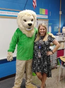 A woman stands smiling in a colorful classroom beside a lion mascot. The mascot wears a bright green shirt and has one arm around her shoulder. She wears a black patterned dress and a lanyard with an ID badge. A whiteboard, number line, and American flag are visible on the blue classroom walls behind them.