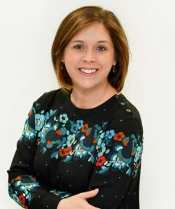 Headshot of Pamela Stepko - A woman with short light-brown hair smiles at the camera against a plain white background. She wears a black top decorated with teal, blue, and red floral patterns. Her arms are loosely crossed as she poses in a relaxed, friendly manner.