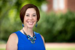 Sarah Tanner Anderson 2020 - A woman smiles while standing outdoors in front of soft green foliage. She wears a bright blue sleeveless top with matching blue and green statement jewelry. Her short brown hair is neatly styled, and she looks directly at the camera. The background is blurred, highlighting her as the focal point of the image.