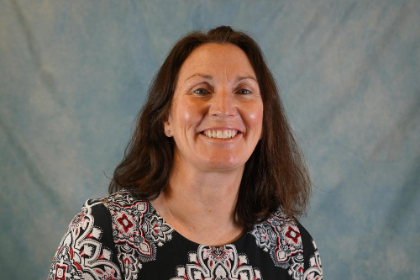 Missy Shores, a woman with long brown hair, smiling in front of a soft blue background. She is wearing a black top with a red, white, and gray patterned design.