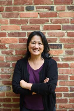 A woman with shoulder-length dark hair smiles with arms crossed, standing against a red brick wall. She wears a purple shirt and black cardigan.