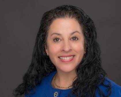 Headshot of Ginny Gills - A woman with long, curly dark hair smiles at the camera against a neutral gray background. She wears a bright blue blazer with a gold clasp and small gold earrings. A delicate gold necklace rests at her collarbone. Her expression is warm and professional.