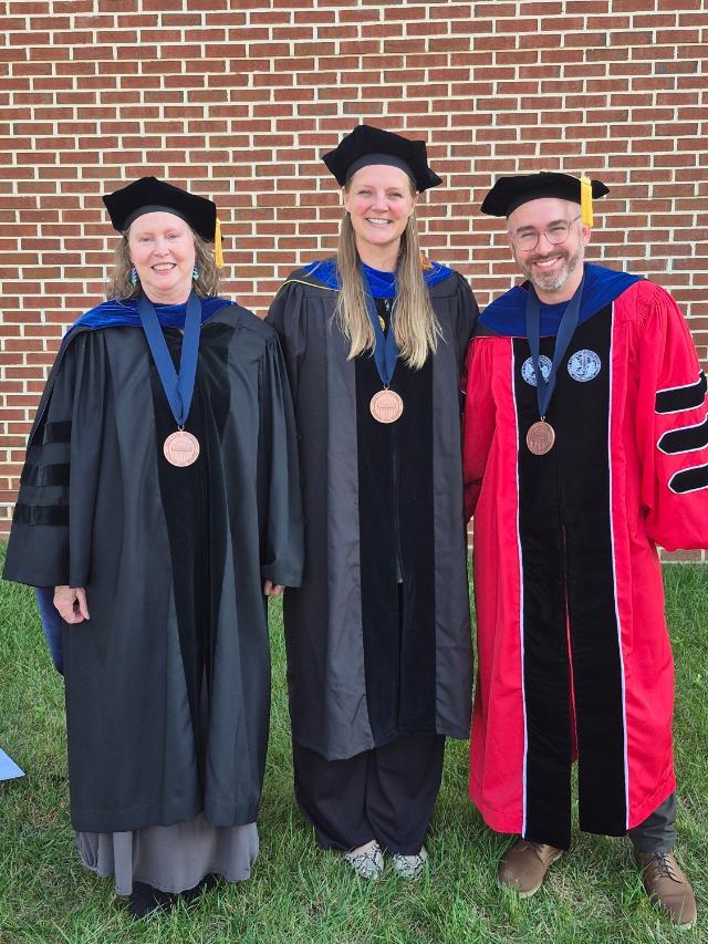 Education and Counseling Faculty Award Winners - Three faculty members in academic regalia stand smiling on grass in front of a brick wall. Each wears a doctoral gown and cap with a medallion hanging on a blue ribbon. The person on the left wears a black gown with velvet panels, the center person wears a black and gray gown, and the person on the right wears a red and black gown. All three face th