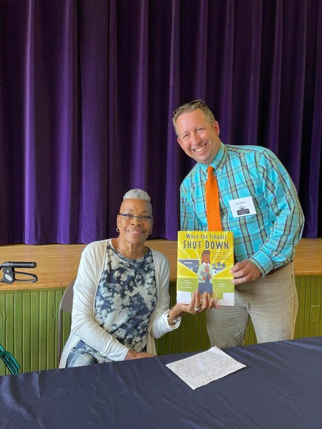 Carl Harvey and Yolanda Gladden, author of When the Schools Shut Down - Two adults smile while holding a children’s book titled *When the Schools Shut Down*. The woman sits at a table, wearing glasses and a patterned top. The man stands beside her in a plaid shirt with an orange tie and name badge. They are in front of a stage with deep purple curtains.
