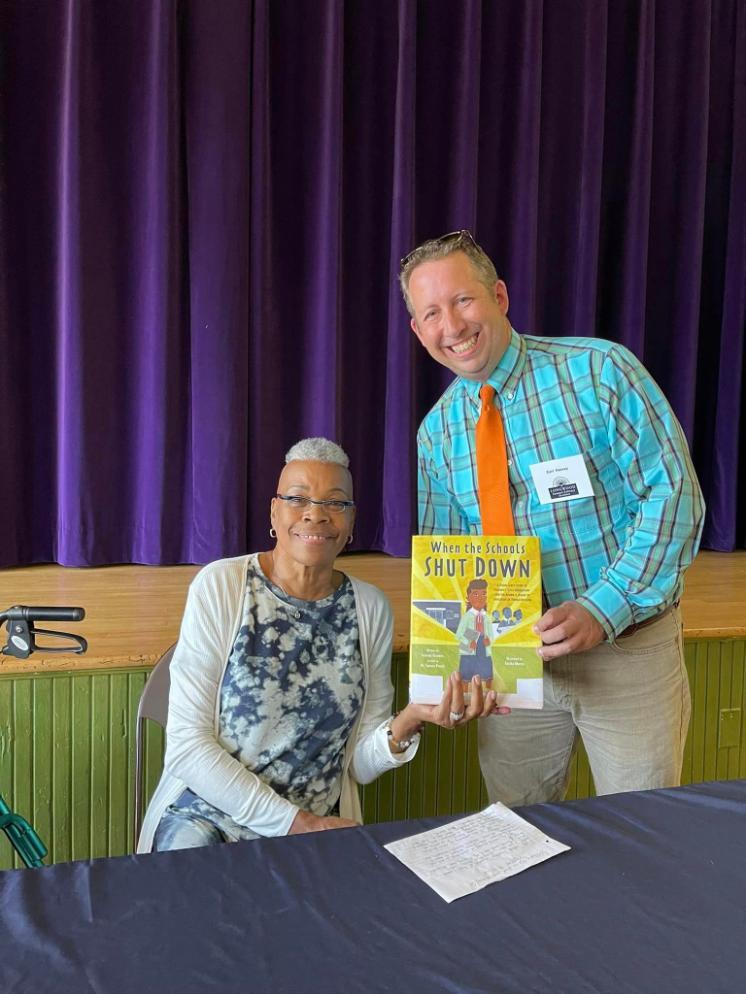 Carl Harvey and Yolanda Gladden, author of When the Schools Shut Down - Two adults smile while holding a children’s book titled *When the Schools Shut Down*. The woman sits at a table, wearing glasses and a patterned top. The man stands beside her in a plaid shirt with an orange tie and name badge. They are in front of a stage with deep purple curtains.