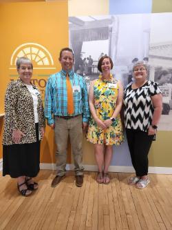 2022 Summer Literacy Institute Planning Committee - Four adults stand smiling in front of a colorful wall display at a museum or educational center. The group includes two women on the ends and one woman and one man in the middle, all wearing name badges. They stand on a light wood floor with a backdrop featuring historic photos and a yellow panel with a window-shaped logo. Each person is dressed