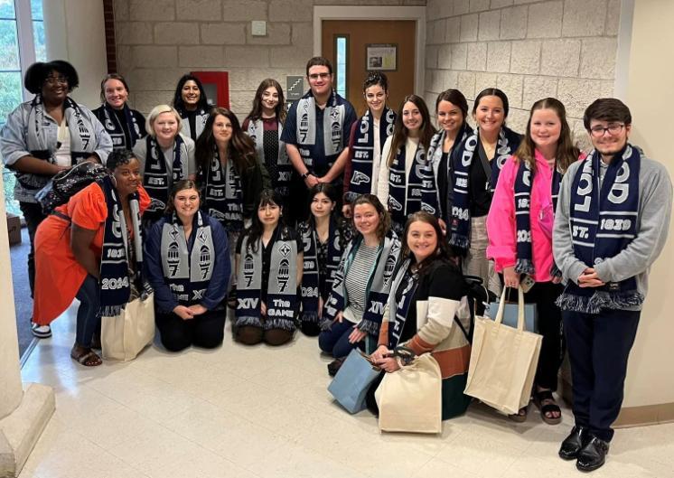 Martinsville students in their Longwood scarves - A large group of students poses indoors wearing matching navy-and-gray Longwood scarves. Some students stand while others kneel in front, smiling at the camera. Several hold tote bags and jackets, gathered in a hallway with brick and beige walls. A door and stairwell sign are visible behind the group.