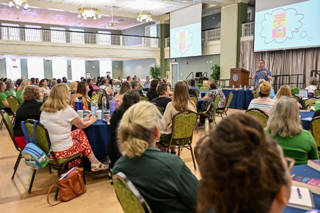 Participants listening to keynote speaker John Schu - A large group of attendees sits at round tables in a bright ballroom. A speaker stands at a podium on stage, presenting with colorful book graphics on two large screens. Participants face forward, listening and taking notes during the session. Chandeliers, high ceilings, and balcony railings add to the open, formal setting.