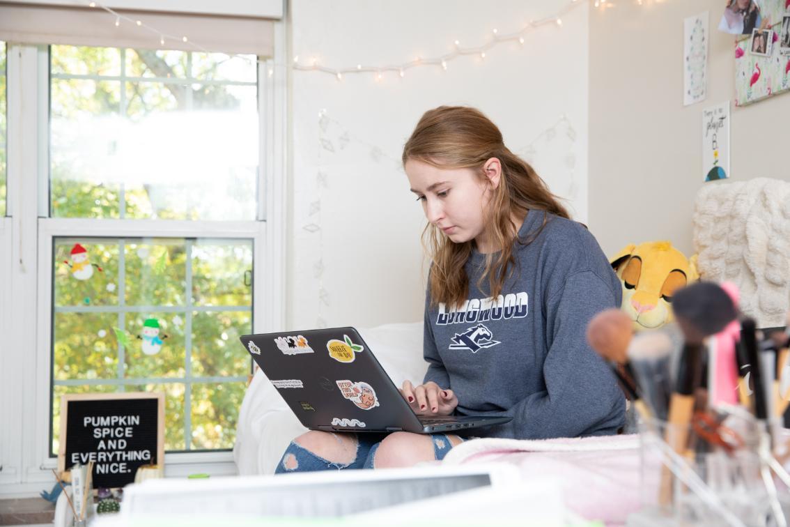 Student working on a laptop sitting on the bed in her residence hall room