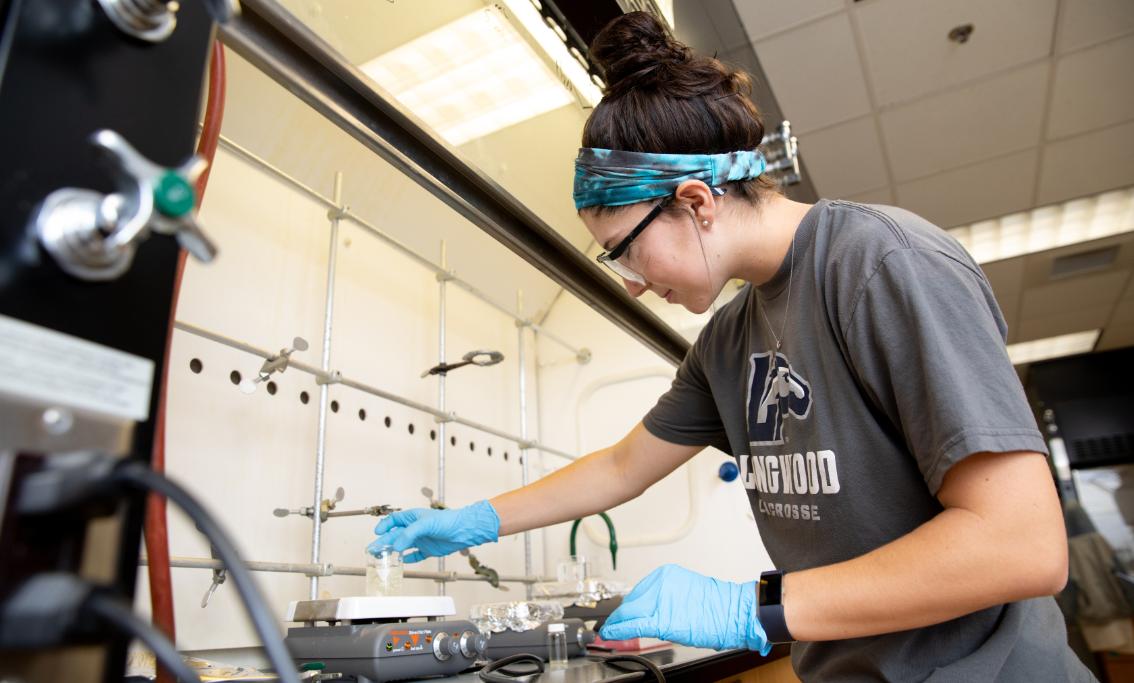 Student working in a research lab - A student wearing gloves and goggles works carefully at a lab bench. She handles a beaker under a fume hood with equipment and tools around her. Bright overhead lighting illuminates the scientific workspace. The scene shows hands-on laboratory learning in a campus science facility.