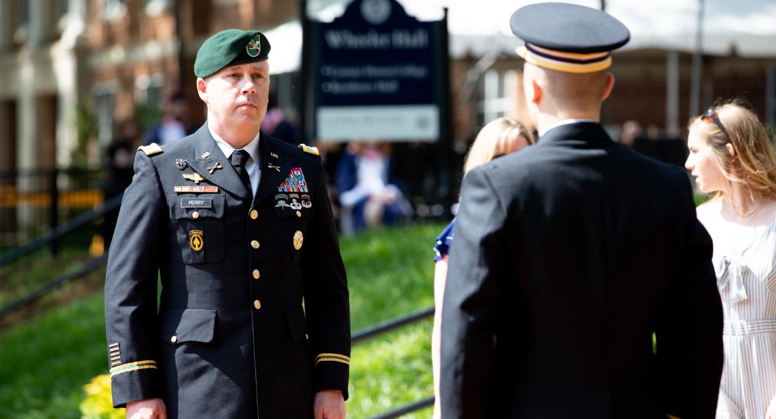 Pinning ceremony for students at graduation - A uniformed military officer stands outdoors during a formal ceremony. He faces another service member whose back is to the camera. Sunlight highlights their dress uniforms against a blurred building and sign. People and greenery in the background suggest a campus event setting.