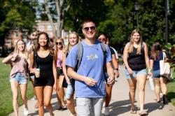 Students walk on campus in a group. The person out front is wearing a blue 3OA Beach Beach Happy shirt, aviator sunglasses and is smiling. - Group of Longwood University students walking together on campus. A student wearing a backpack smiles at the front of the group. Students wear casual clothing and carry drinks and bags.