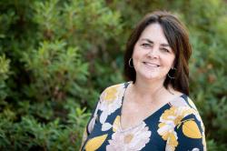 Headshot of Caroline Gibbs - Portrait of a woman smiling outdoors on Longwood University’s campus. She wears a patterned blouse with floral designs in yellow and cream. Dark shoulder-length hair and hoop earrings frame her face. Green foliage creates a soft, natural background.