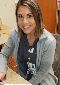 2018 Alumnae Darcy King, Therapeutic Recreation - A smiling woman sits at a desk in an office setting. She wears a gray zip-up jacket and an ID badge clipped to her shirt. Her shoulder-length hair is highlighted and neatly styled. Office cabinets, a countertop, and work materials are visible around her.