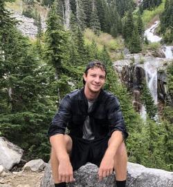 Head shot of Nate Wilson, 2018 Alumni  Therapeutic Recreation - A man sits on a rock outdoors, smiling at the camera. He wears a dark long-sleeve shirt and black shorts. Evergreen trees and a rocky mountainside surround him. A waterfall cascades down the slope in the background.