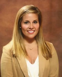 Ellen Sweat Turner ’17 - A professional headshot of a woman smiling toward the camera. She has long blonde hair and wears a tan blazer over a white top. A simple necklace and stud earrings complement her attire. The background is a neutral studio backdrop with warm tones.
