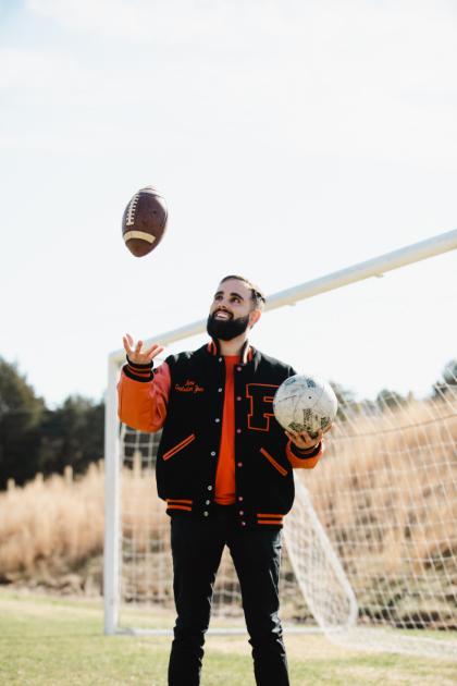 A man standing on a grassy field in front of a soccer goal, wearing a black and orange varsity jacket with a large letter