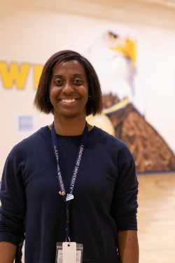 - A smiling woman stands indoors wearing a Longwood University lanyard. She wears a dark sweater and an ID badge around her neck. Her hair is cut to shoulder length and parted naturally. A softly blurred mural fills the background behind her.