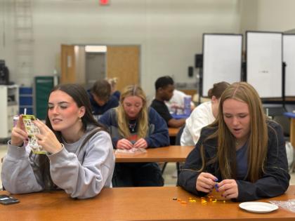 Students participate in LEGO activity in KINS 205 - Students sit at tables working on a hands-on classroom activity. Two students in the foreground sort small colorful items and read instructions. Additional students focus on similar tasks in the background. The setting appears to be an active learning lab or classroom environment.