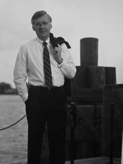 Photo of Governor Balilies. - Black-and-white photograph of a man standing near a waterfront. He wears a dress shirt, tie, and glasses, with a jacket draped over his shoulder. Wooden pilings and chain barriers appear behind him along the water’s edge. The image has a formal, reflective tone with a coastal setting.