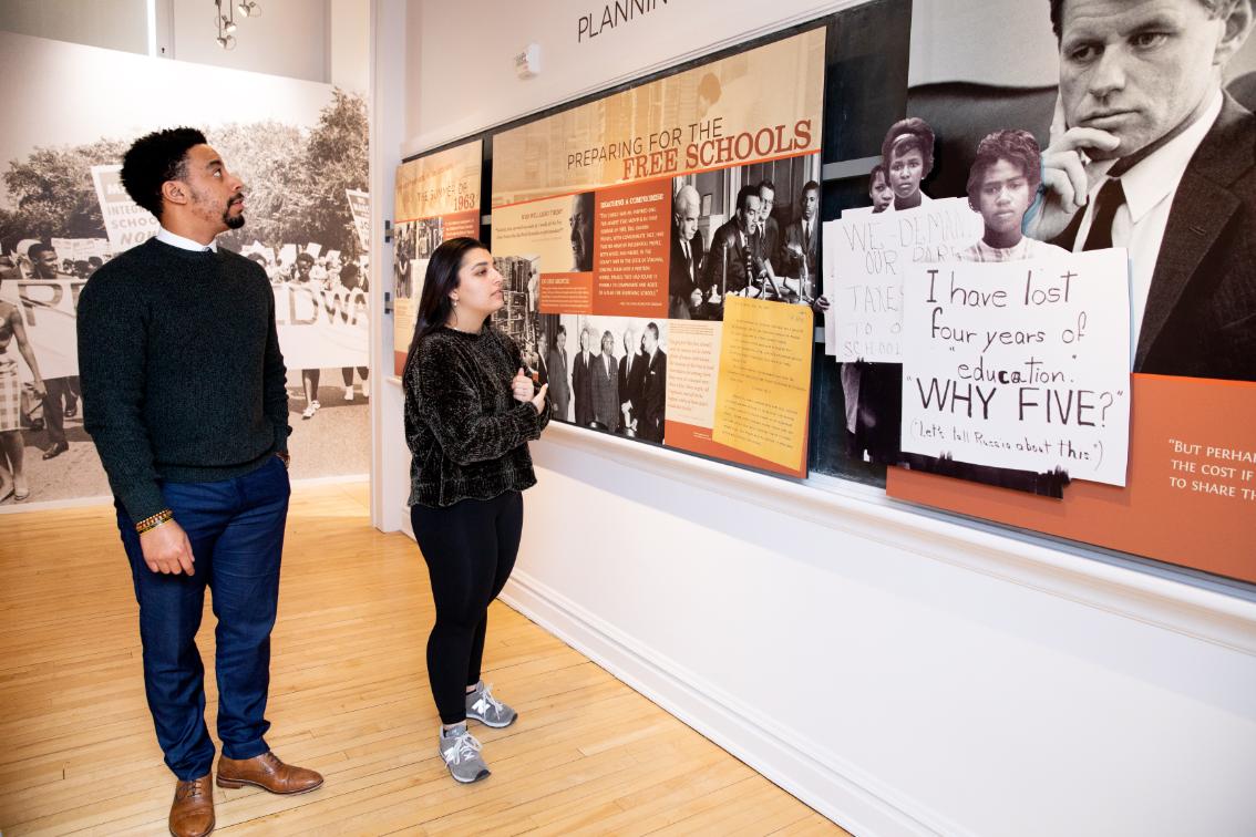 Visitors at the Moton Museum - Two people stand in a museum gallery looking at a civil rights exhibit. Large black-and-white photos and protest signs cover the wall display. The exhibit includes panels titled “Preparing for the Free Schools.” Wood floors and bright lighting frame the exhibit space.