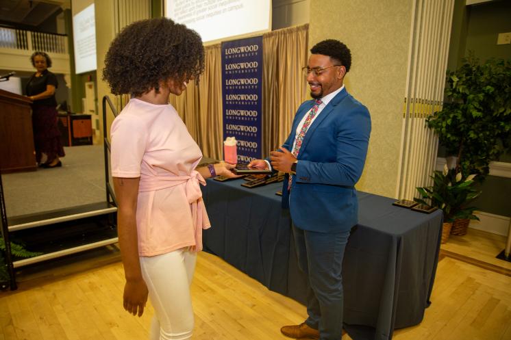 A student receives an award from a presenter during a ceremony. They smile as the presenter hands over a plaque on stage. Longwood University banners hang behind them. An audience member and podium are visible in the background.