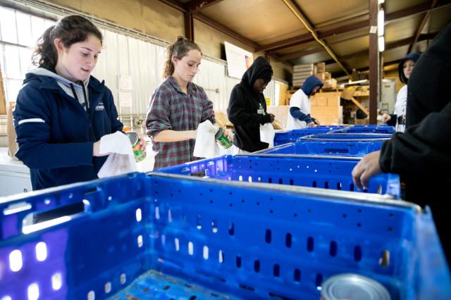 Students line up at a warehouse-style facility cleaning canned goods. They use white cloths to wipe the cans over large blue bins. The group works together in a bright, industrial setting. Stacks of boxes and supplies fill the background.