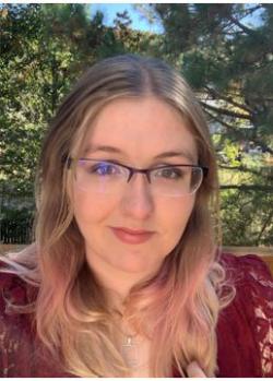 heather switzer - Portrait of a woman with long blonde hair wearing glasses. She is dressed in a maroon top and a necklace. The photo is taken outdoors with trees and greenery in the background. The image shows her head and shoulders facing the camera.