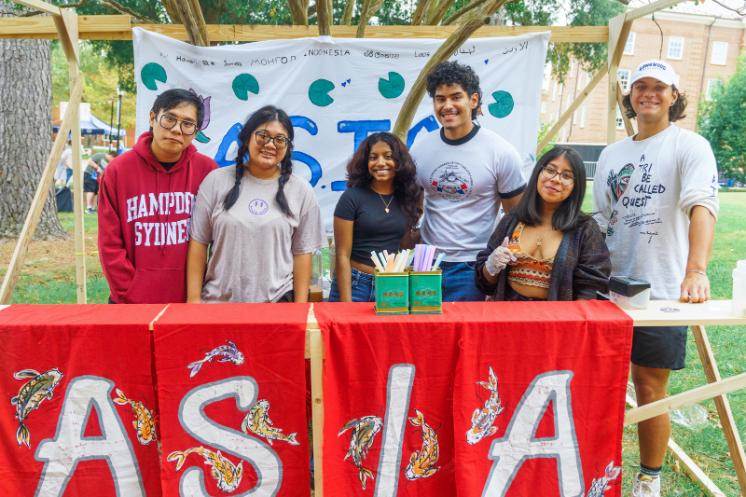 ASIA booth at Oktoberfest 2023 - A group of students stands behind a table at an outdoor campus event booth. A red banner reading “ASIA” hangs across the front of the table. They smile at the camera with supplies and decorations arranged in front of them. A handmade backdrop and campus buildings are visible behind the group.