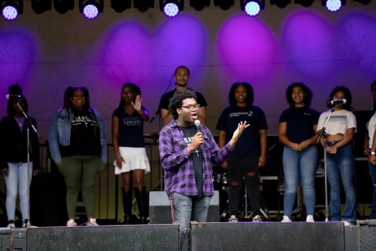 BASIC Gospel Choir performing at Oktoberfest - A student performs on stage holding a microphone and singing. A group of singers stands behind as backup performers. Purple stage lighting illuminates the performance area. Microphones and musical equipment are visible on stage.